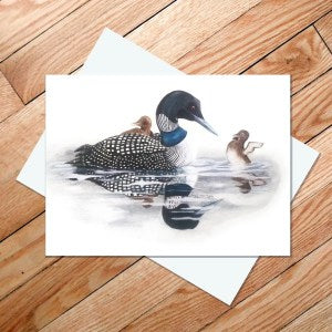 Illustration from the "Great Northern Loon" 5x7 card, depicting a Common Loon swimming with two ducklings on its back, set against a wooden floor background.