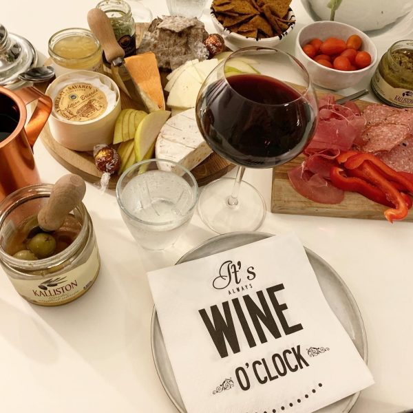 A wine lover's dream spread graces the table, featuring a mix of cheeses, cured meats, sliced red peppers, cherry tomatoes, olives, and bread. A glass of red wine sits nearby with eco-labeled "Napkins: It's Always Wine O'Clock" adding the perfect touch.