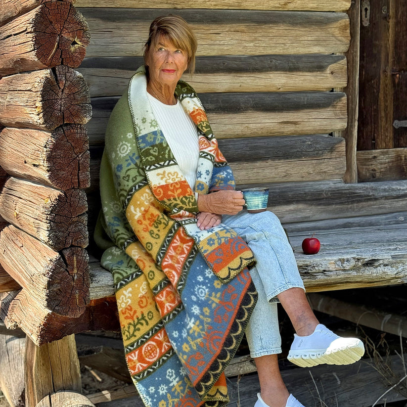 A woman sits on a wooden porch wrapped in a wool blanket with a colorful, Nordic-inspired patchwork pattern.