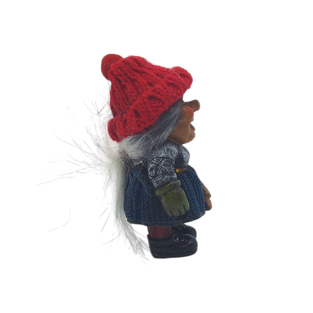 A Scandinavian-inspired Nisse figurine sports a red knit hat and gray hair, with a greenish face. The side view reveals a gray top, blue patterned skirt, and black shoes, capturing the essence of a traditional Nisse wife.