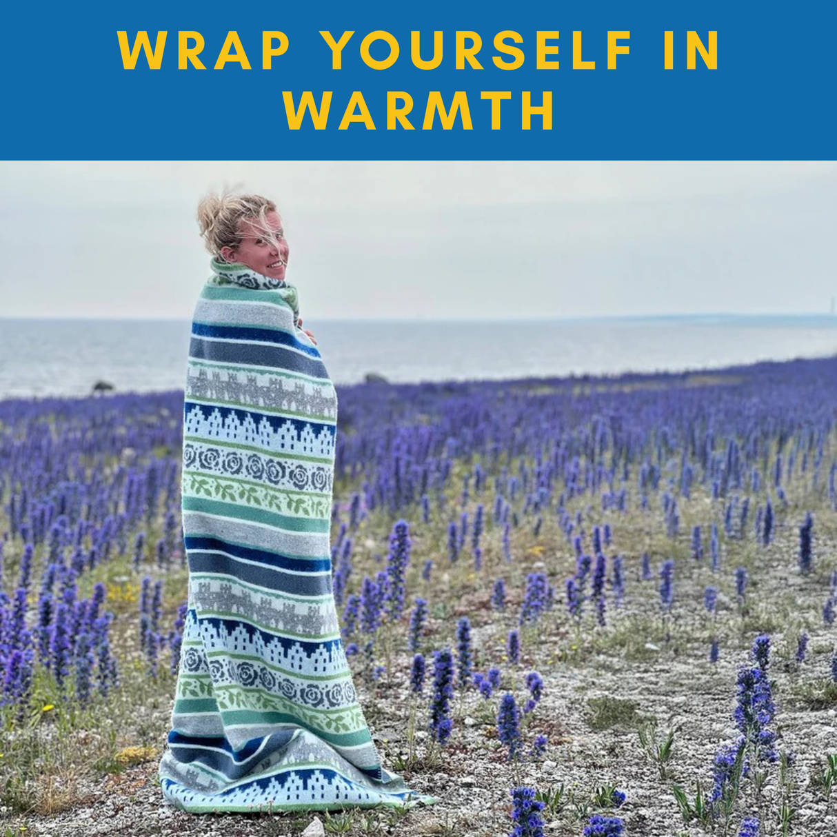 A person wrapped in the Black Dala Horse Pattern Dalarna Wool Blanket (100% Norwegian Lambswool, 130×180 cm) stands in a field by the sea under clouds. Swedish design on display; text above reads, Wrap yourself in warmth.