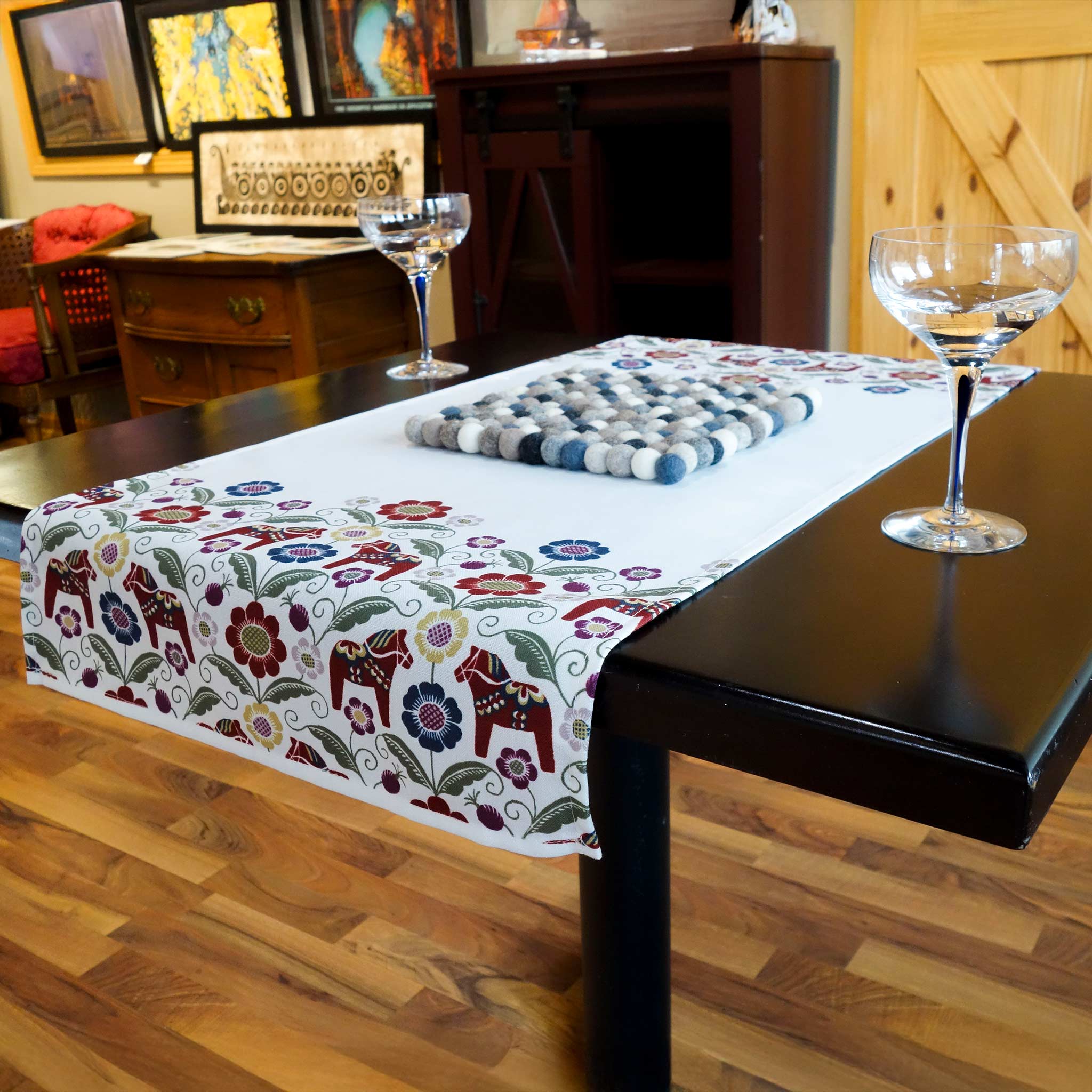 A black table with the Siljan Dala-inspired runner, featuring Dala horses, flowers, and kurbits, holds two empty wine glasses on a decorative wool mat. Its set against a wooden floor amid art and furniture that enhance its elegance.