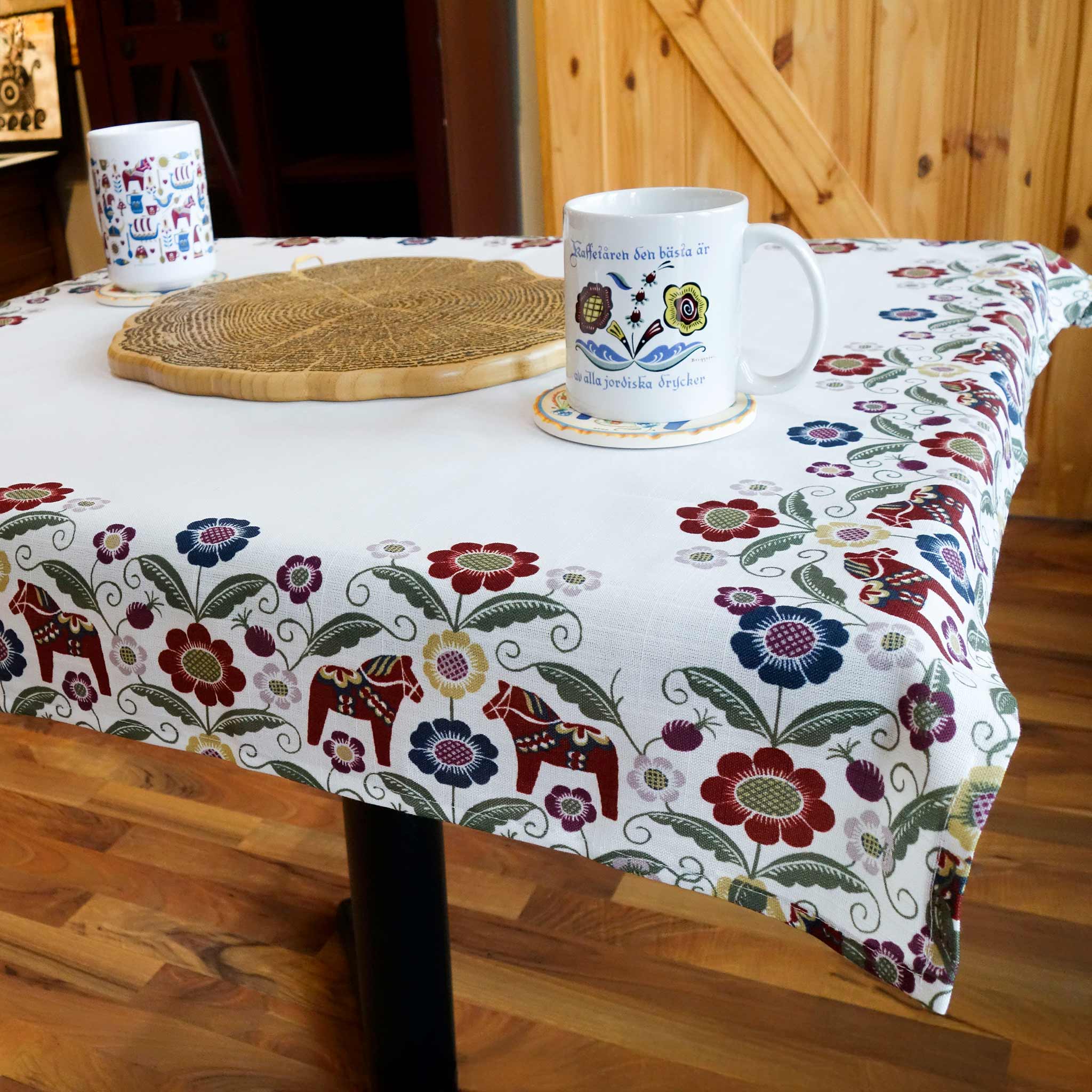 A square table with a Siljan Tablecloth decorated with Dala horses, flowers, and Kurbits designs, two white mugs, and a woven placemat in a warm wooden interior.