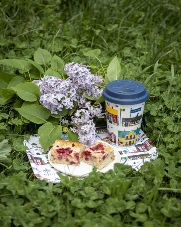 A Sweden To Go porcelain travel mug is arranged on a napkin in the grass with two pieces of berry cake and lilac flowers.