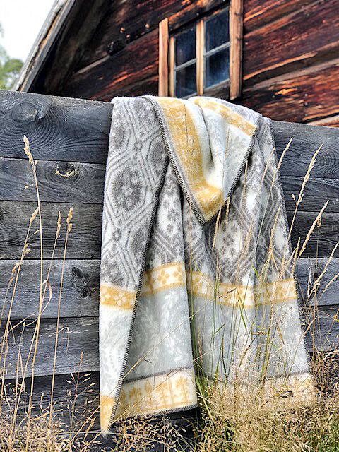 A Brunkulla Cornflower Brown blanket by Kerstin Landstrom, crafted from 100% Norwegian lambs wool, featuring grey and yellow designs, drapes over a wooden fence with an old wooden building in the background.
