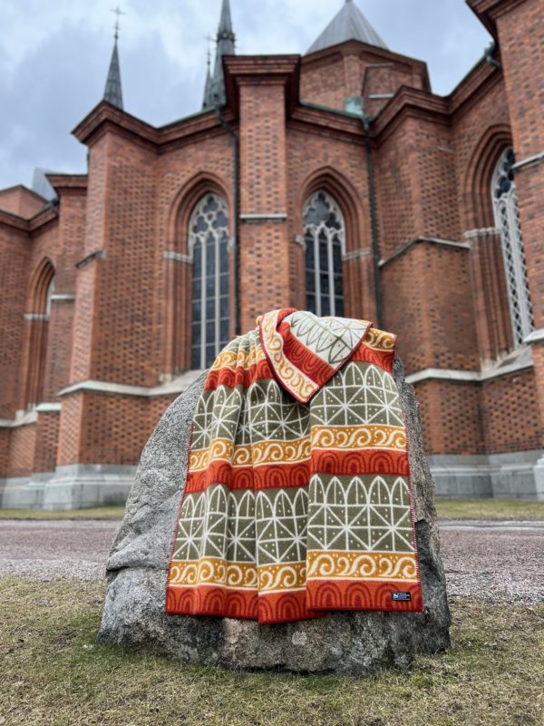 An Uppsala blanket, made of 100% Norwegian lambs wool by Kerstin Landstrom, is draped over a rock in front of a brick church with tall spires and arched windows. With its architectural pattern and OEKO-TEX certification, its an ideal graduation gift.