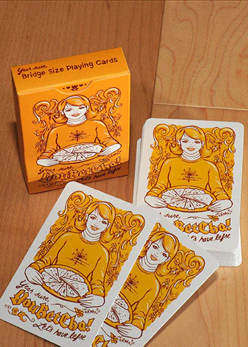 A box of Hotdish/YouBetCha Bridge Size Playing Cards, featuring three face cards illustrated by Adam Turman with a woman in an orange shirt holding a pie and vibrant orange decorative designs.