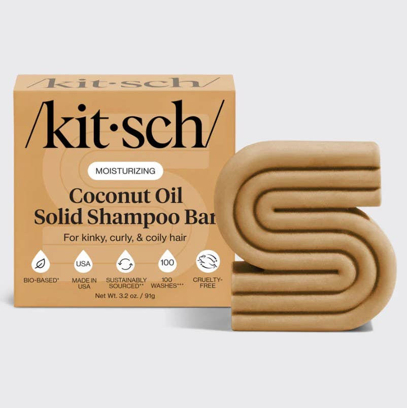 The KITSCH Coconut Oil Shampoo Bar for Dry Damaged Hair (3.2 oz) is uniquely designed in an abstract “S” shape and comes next to its tan packaging. Specially labeled as a moisturizing shampoo for kinky, curly, and coily hair, this solid bar highlights icons for bio-based ingredients, USA-made quality, cruelty-free standards, and sustainable sourcing. Infused with nourishing coconut oil, it helps enhance and care for your natural curl pattern.