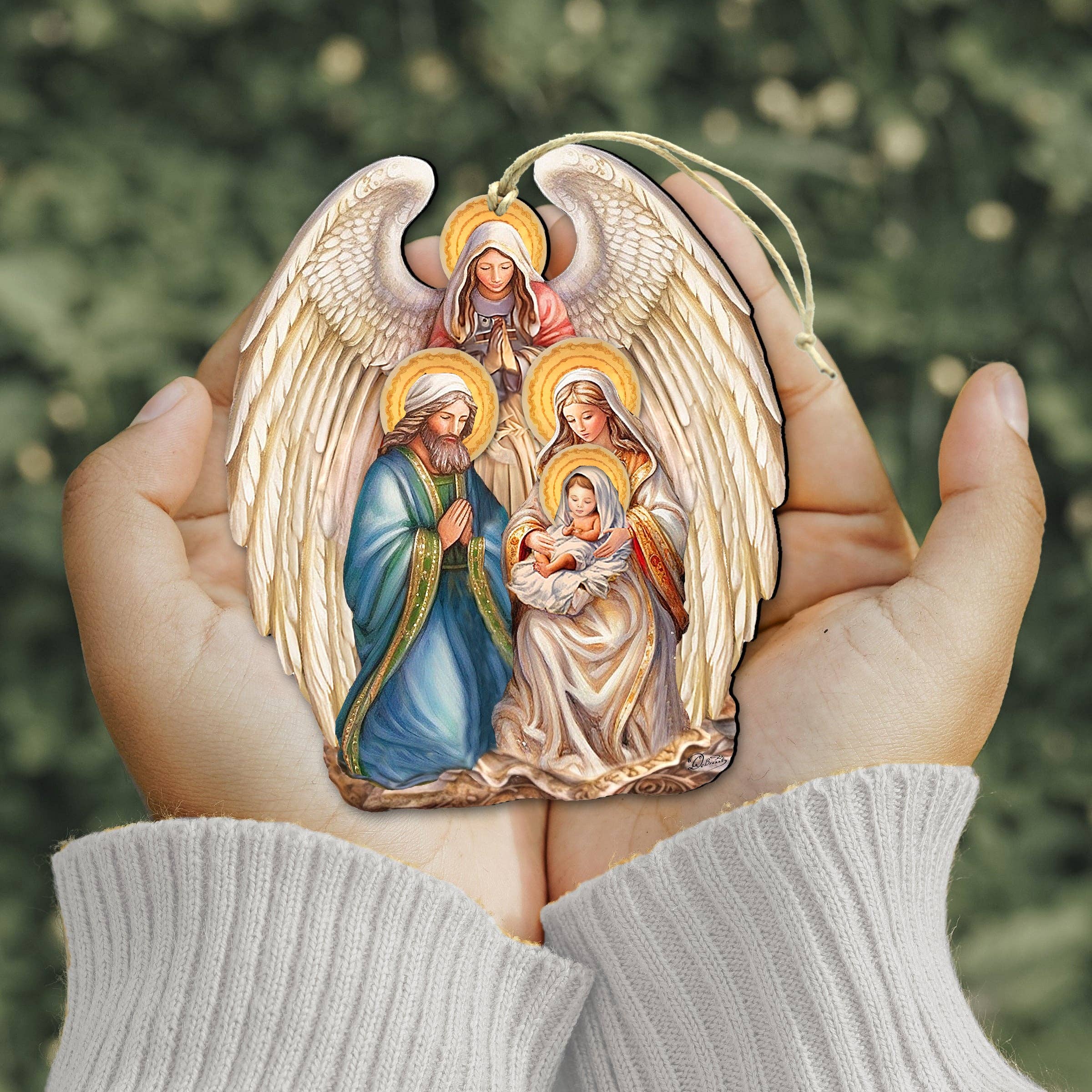 Someone holds the Ornament: Angel with Nativity Family Wooden Christmas Tree Ornament by G.Debrekht (5x4 inches) in front of a blurred green background.