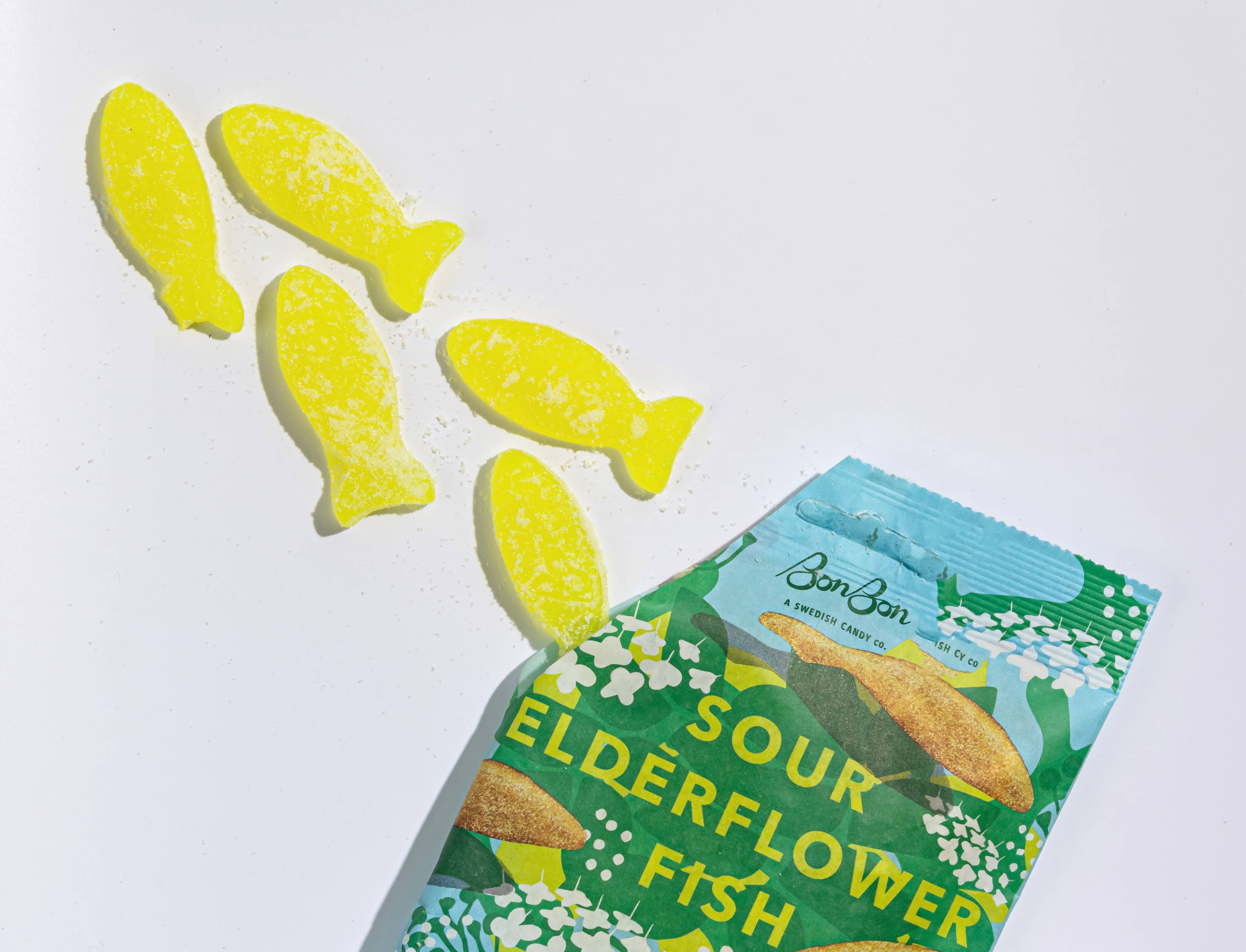 An open bag of BonBon Sour Elderflower Fish (150g) Swedish candy, with four yellow, vegan, gluten-free fish-shaped candies spilling onto a white surface.