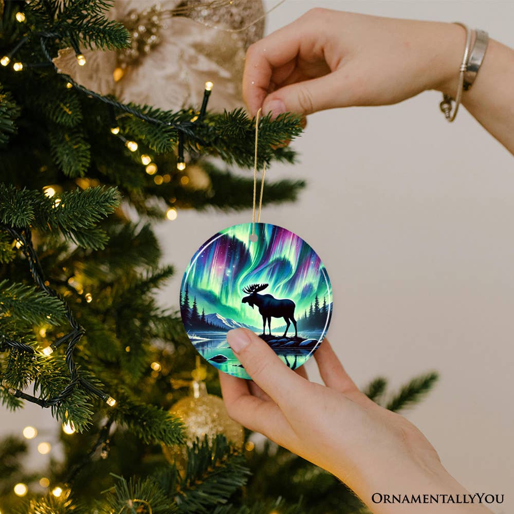 Hands hang the Ornament: Majestic Moose Under Northern Lights Christmas Tree Ornament, featuring a moose silhouette against a Northern Lights background, on a beautifully decorated holiday tree.
