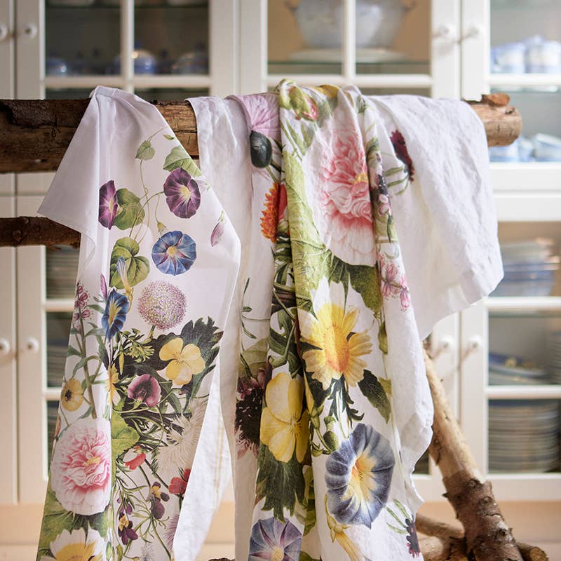 The Tea Towel: A Flower Garden by Jim Lyngvild Koustrup & Co Denmark, made from 100% organic cotton, elegantly drapes over a wooden rod. This charming accessory enhances the kitchen settings style and functionality against cabinets and dishes.