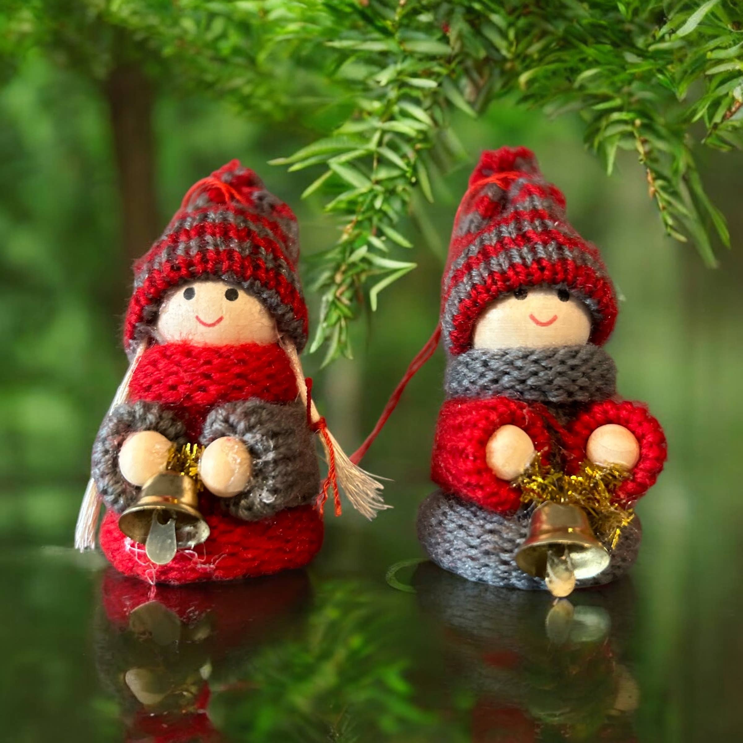 Add a charming Scandinavian touch to your décor with the Ornament: Tomte Boy or Girl with Bells—handcrafted wooden gnomes in red hats and scarves, holding bells, positioned under green foliage.
