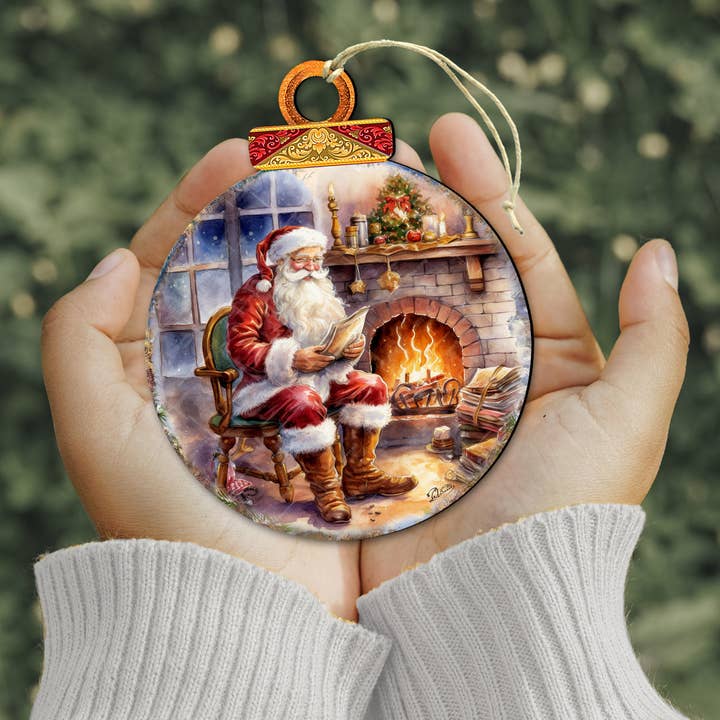 A person holds the Ornament: Santa by the Fireplace Wood Ornament (approx. 4.5" x 4"), which features Santa Claus reading by a festive, holiday-decorated fireplace.
