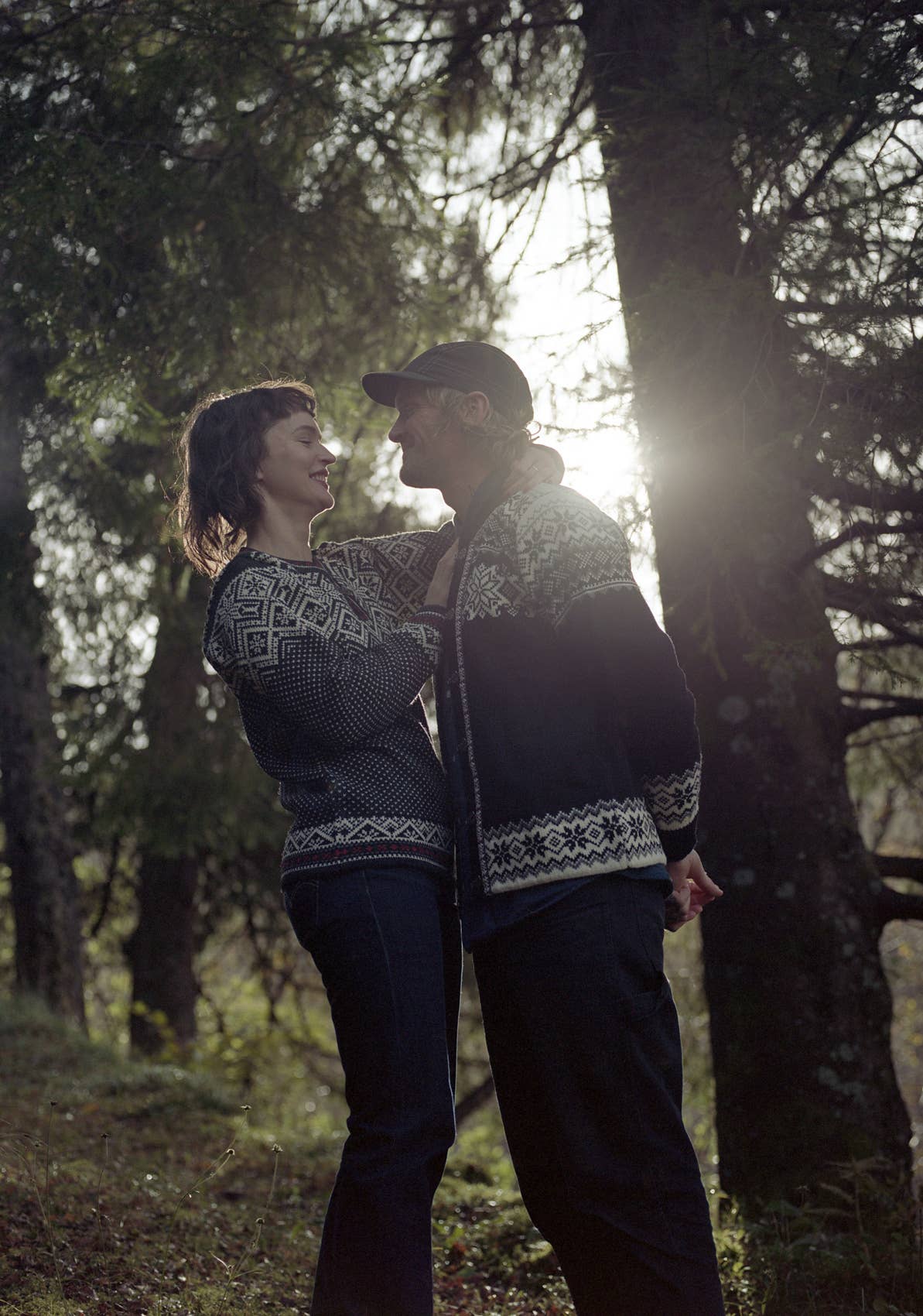 Two people wearing Stavanger Norwegian Wool Cardigan in black stand close together in a forest, sunlight streaming through the trees behind them.