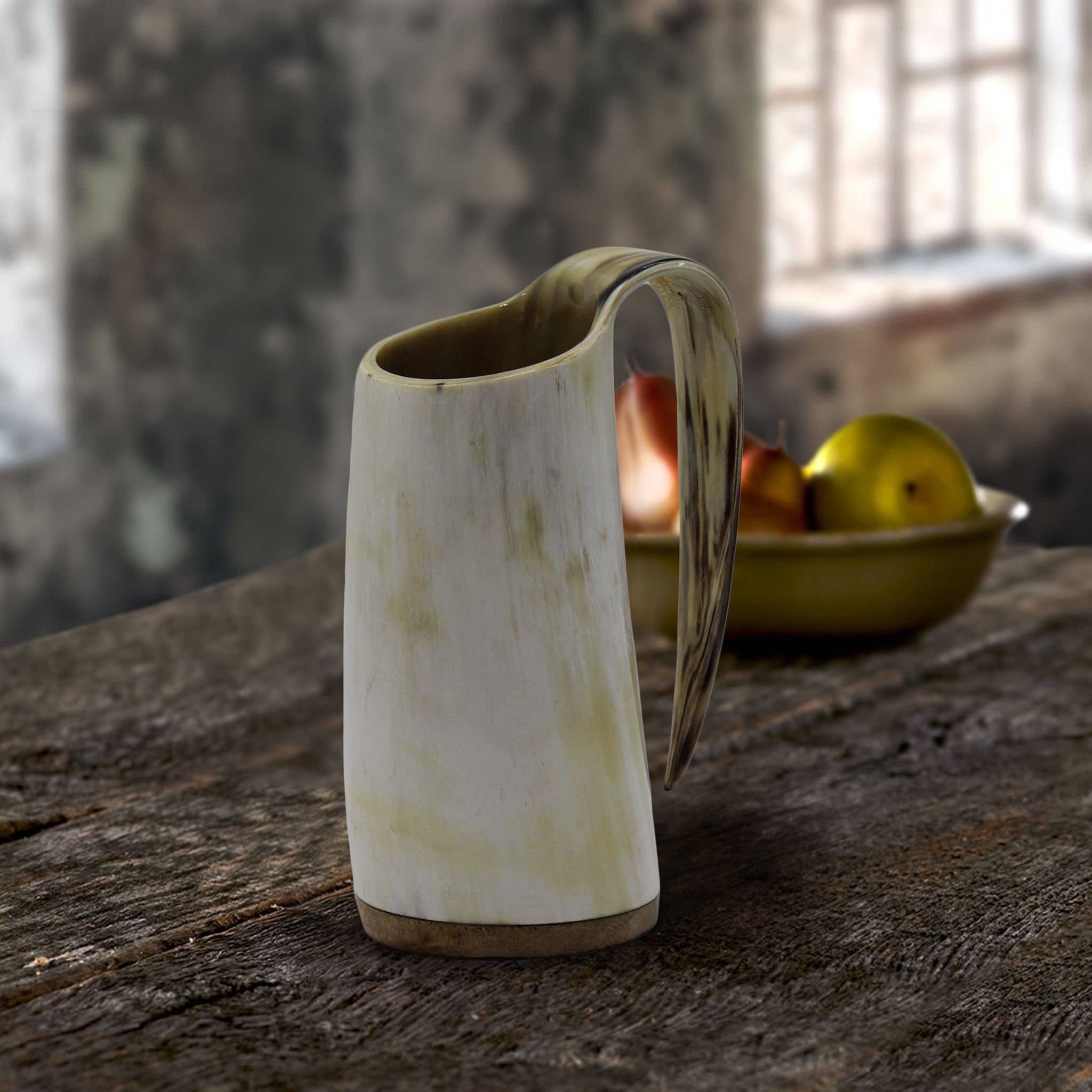 A rustic ceramic pitcher sits on a wooden table, complemented by a blurred backdrop of apples and onions near the window. Among them lies the Horn Mug: Viking African Ox Horn Mug - 100% Authentic Beer Ox Horn Tank, holding 16oz, echoing tales of Viking feasts and hearty gatherings.