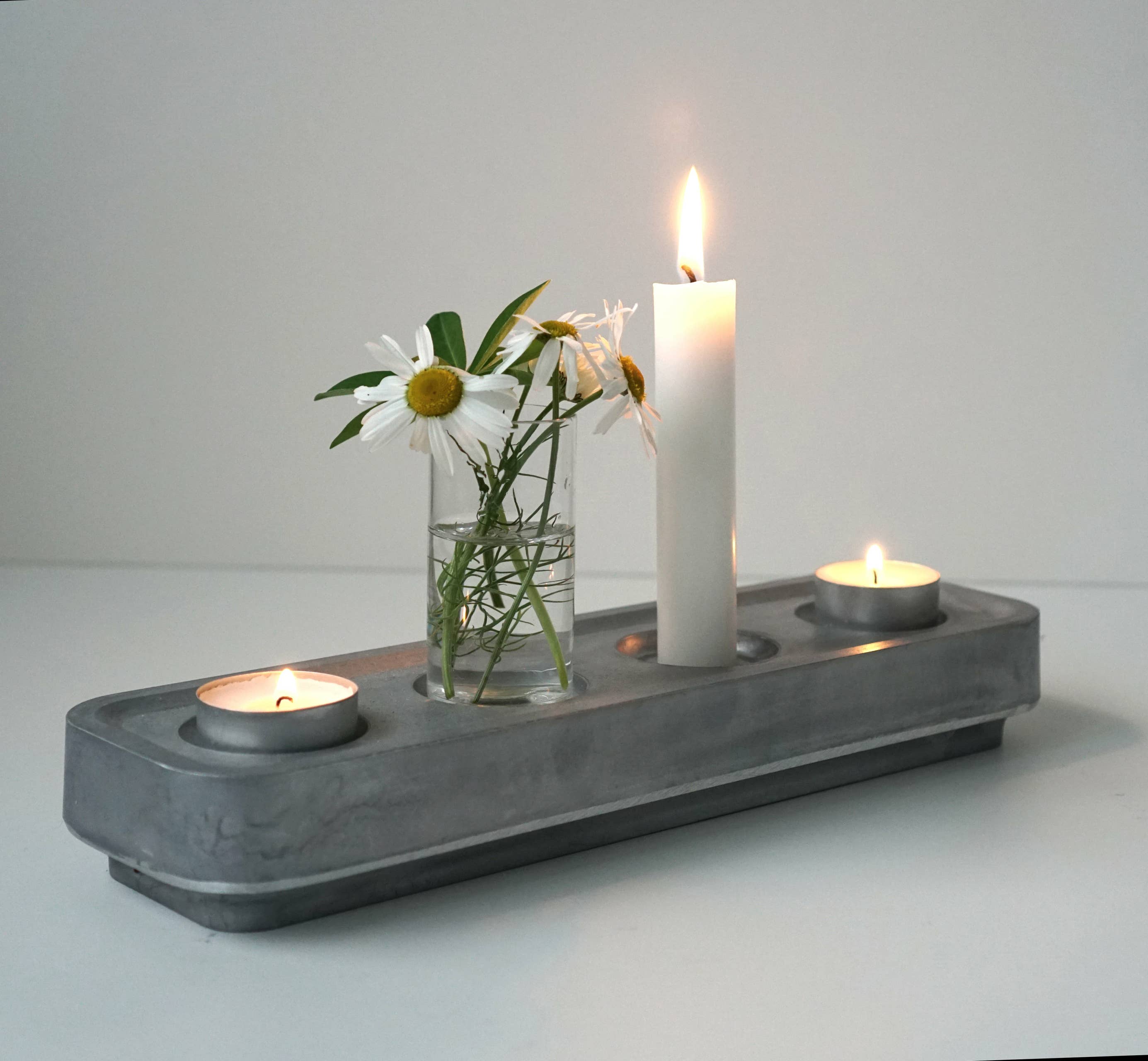 A concrete tray displays two tealight candles, a lit pillar candle, and a clear glass vase with white daisies against a plain backdrop. The scene exudes eco-friendly charm with the Stumpastaken Swedish Candle Holder crafted from recycled materials.