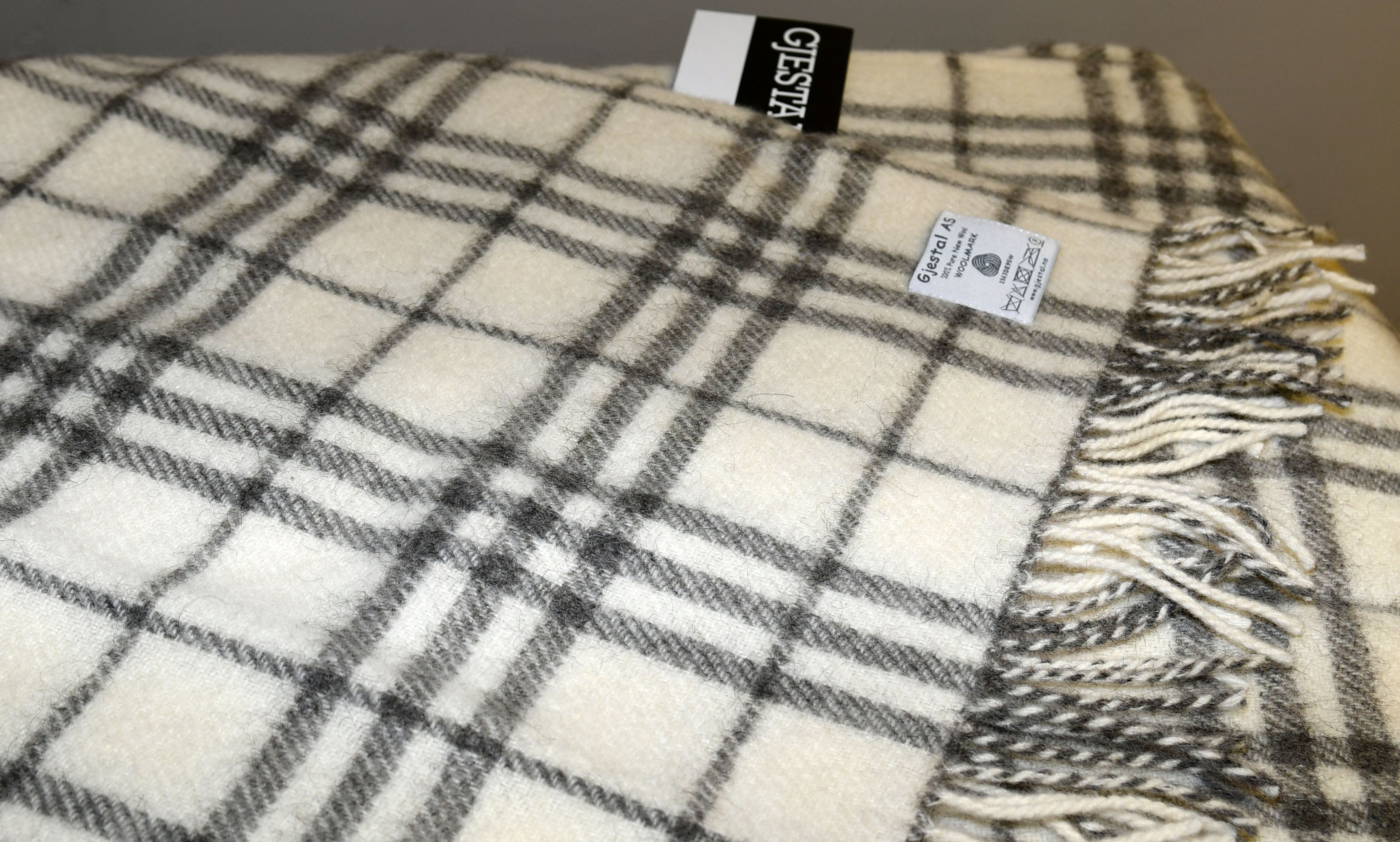A folded Pledd Rutemnster Gjestal AS blanket, featuring a cream and gray plaid pattern with tassels, is shown. It is crafted from Pure New Wool and includes a label that proudly states it is Made in Sweden.