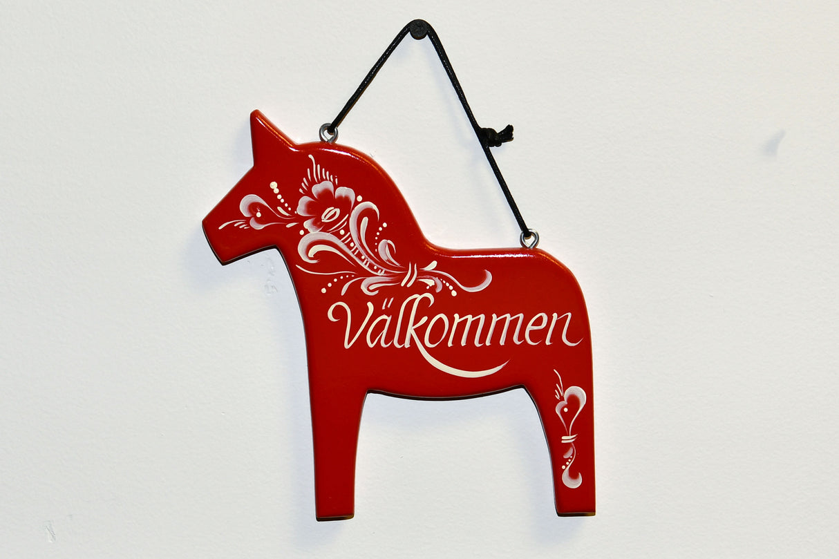 The Välkommen Sign: Swedish Dala Horse Wall Decor features a red, horse-shaped plaque with patterns, crafted from Swedish Pine. It hangs by a black string on a white background, adding charm to any decor.