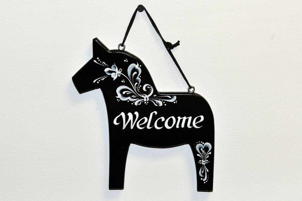 A black Swedish pine Dala horse-shaped Welcome wall decor features hand-painted white floral designs and hangs by a black cord against a white background.