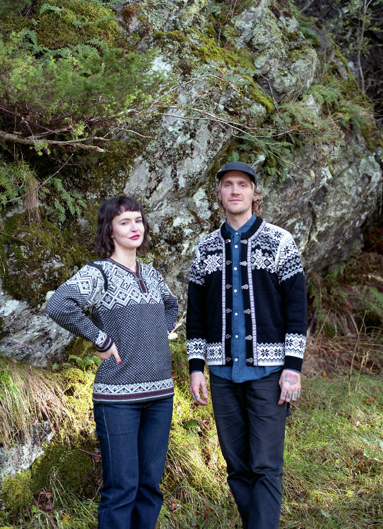 A woman and a man stand outdoors in front of a mossy rock and greenery, wearing the Stavanger Norwegian Wool Cardigan in black.