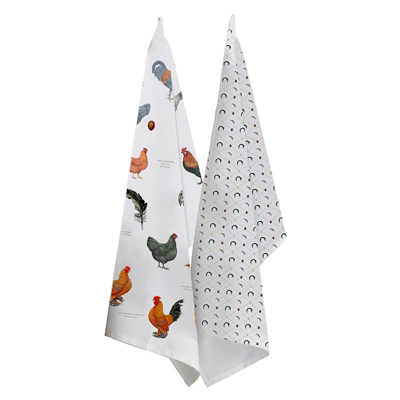 Two kitchen towels hang side by side; one features Annemarie Haase's vibrant rooster illustrations crafted from 100% organic cotton, and the other displays a cheerful pattern of small rainbows.