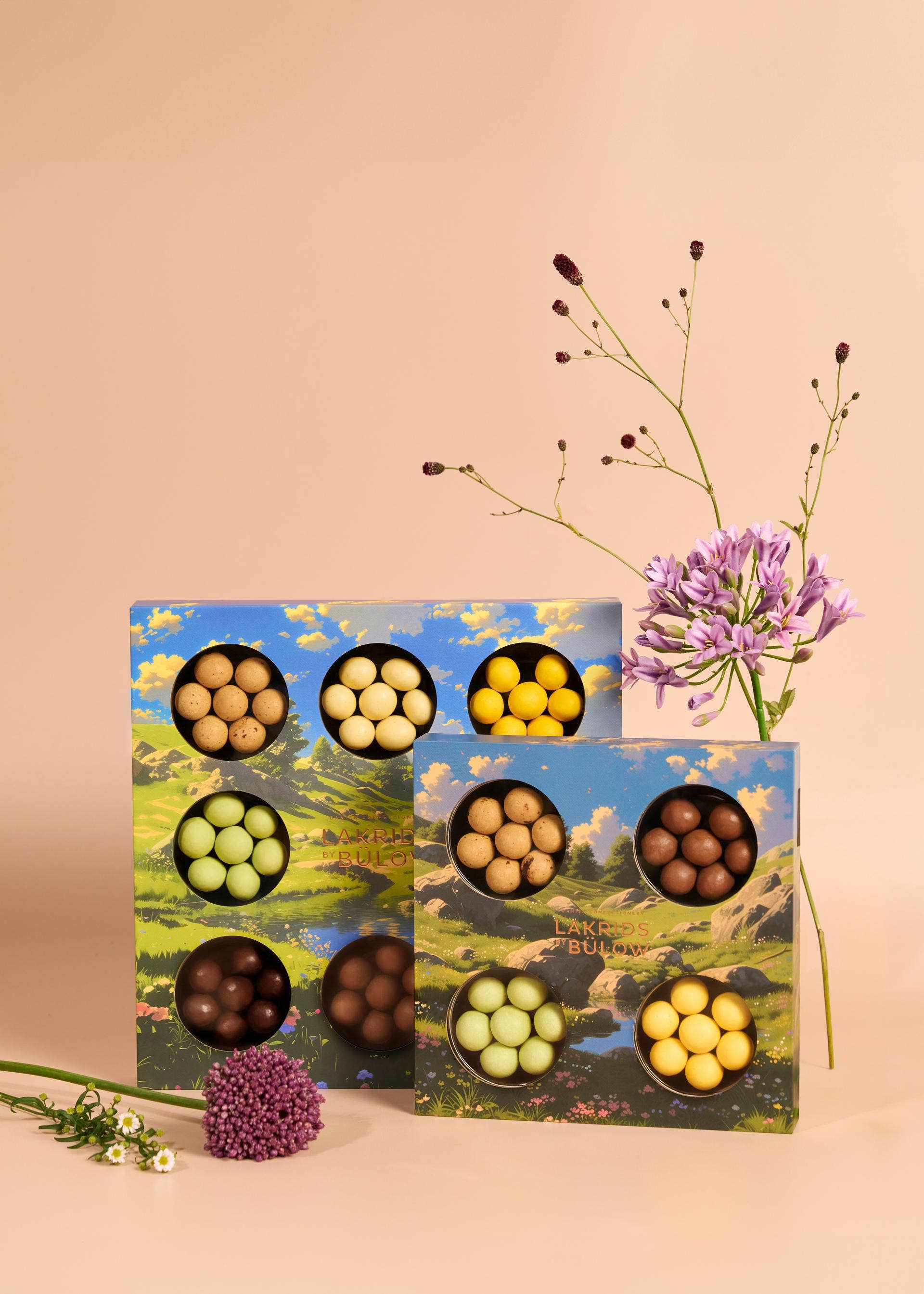 The Lakrids by Bülow SPRING 4-Flavor Gift Box (6.2 oz) features assorted Danish chocolate-coated licorice in colorful boxes with a mountain design, beautifully displayed among purple and white flowers—perfect for gourmet candy lovers.