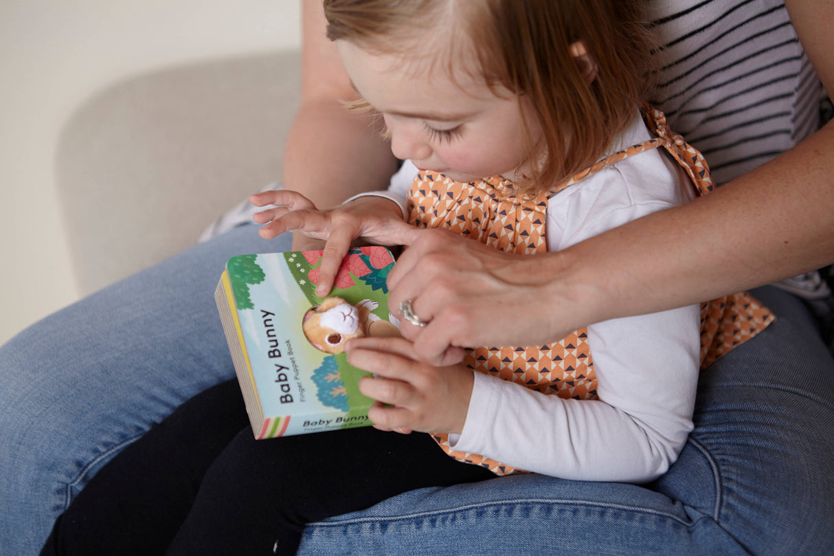 A young child sits on an adult’s lap, exploring the Book: Baby Bunny Finger Puppet Book together as the adult helps turn its interactive pages.