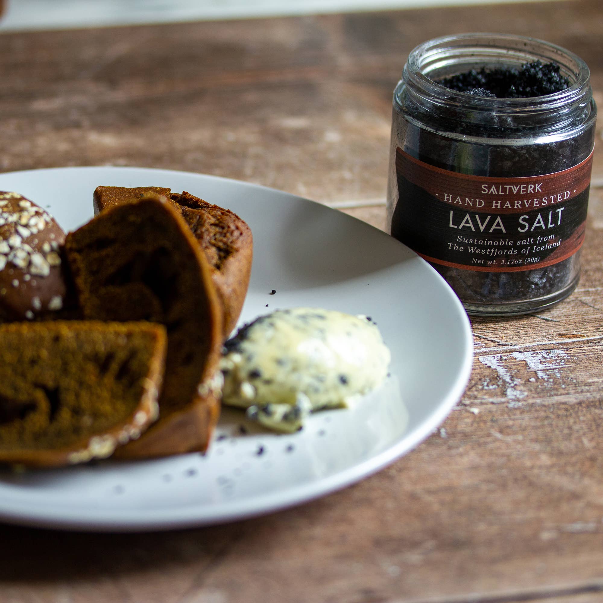 A white plate with dark bread and butter topped with Icelandic Lava Salt from the SALTVERK Gift Set—featuring both Sea Salt & Lava Salt—sits beside a Hand Harvested Lava Salt jar on a wooden table.