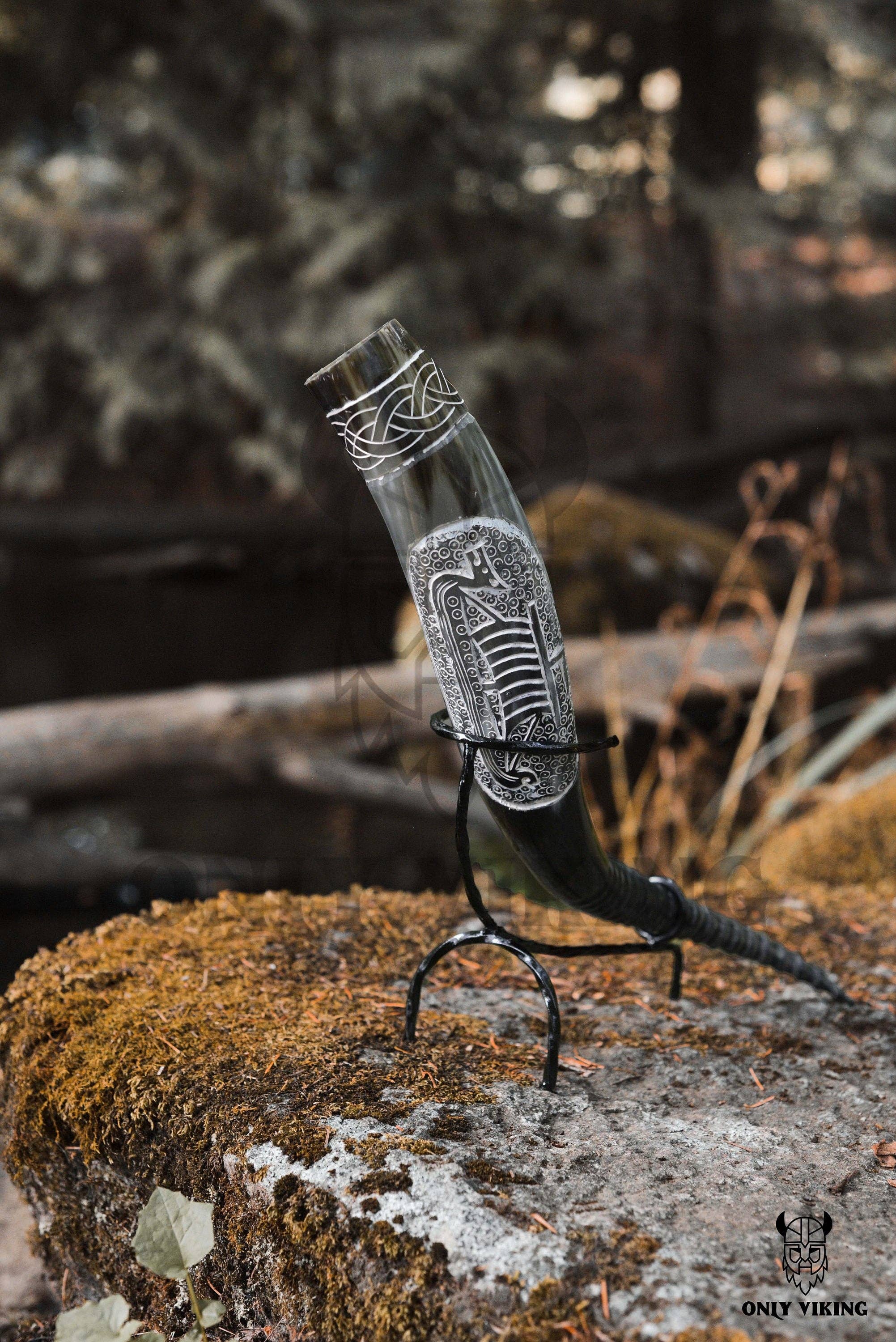 The Ravens Viking Ship Handcrafted Drinking Horn (14-18 inches) stands upright on its metal stand outdoors, resting on mossy stone. Intricate patterns highlight its unique Viking design.