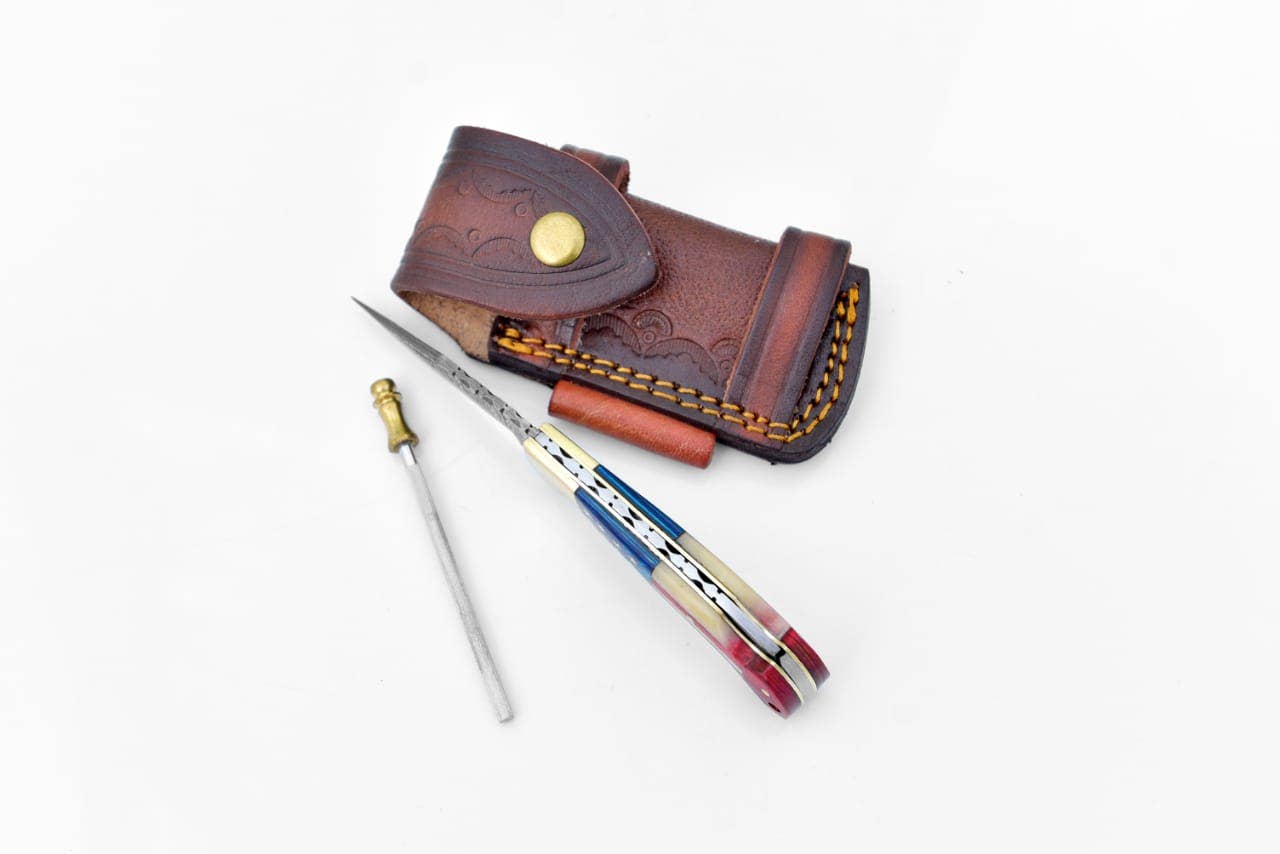 The Knife: Damascus Steel Pocket Knife Titan Patriot features a multicolored handle, narrow blade, and brown leather sheath with yellow stitching—ideal for everyday carry and shown on a white background.