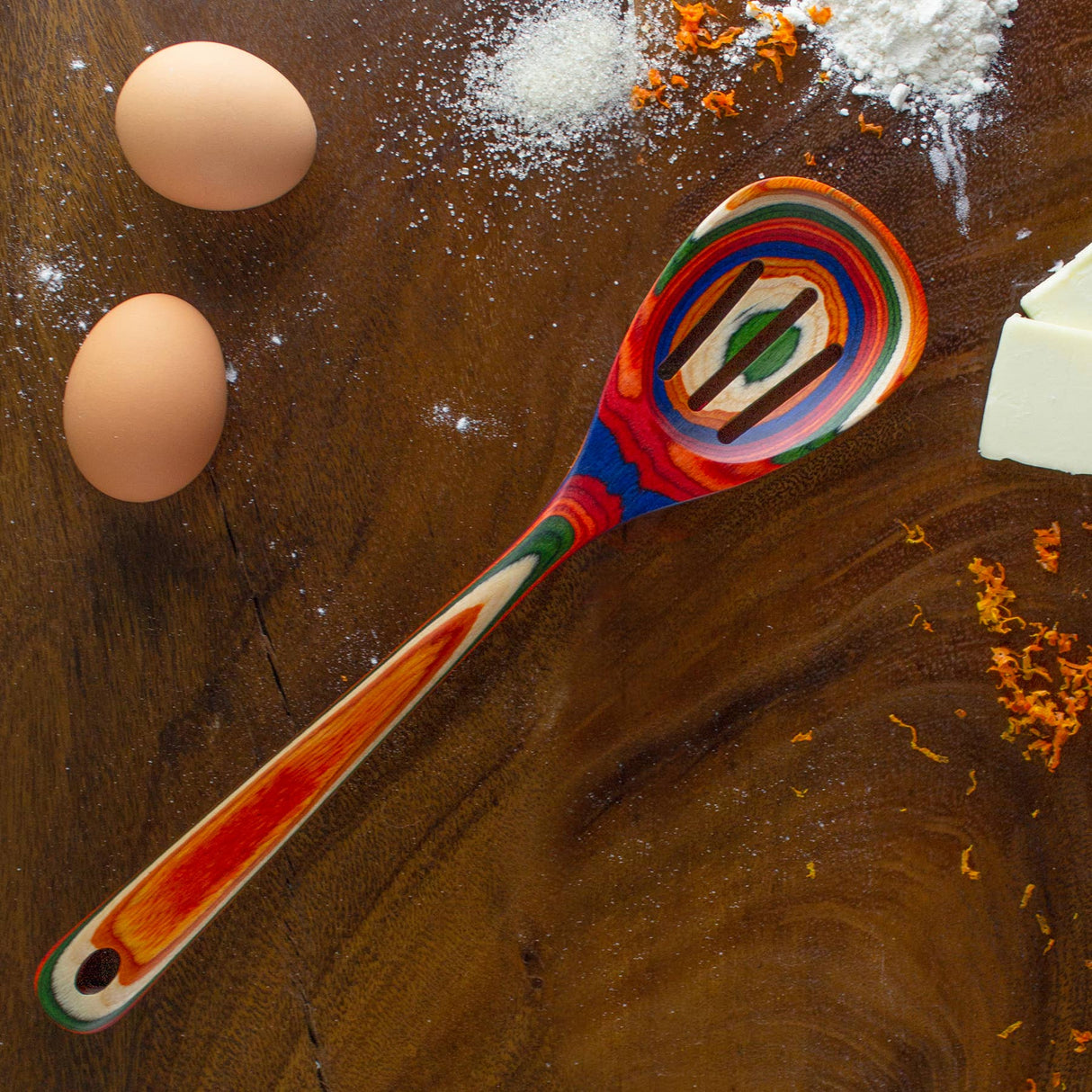 The Marrakesh Collection Slotted Wooden Cooking Spoon lies on a wooden surface surrounded by two eggs, flour, orange zest, and butter. This colorful utensil is non-stick safe for easy cooking and cleaning.