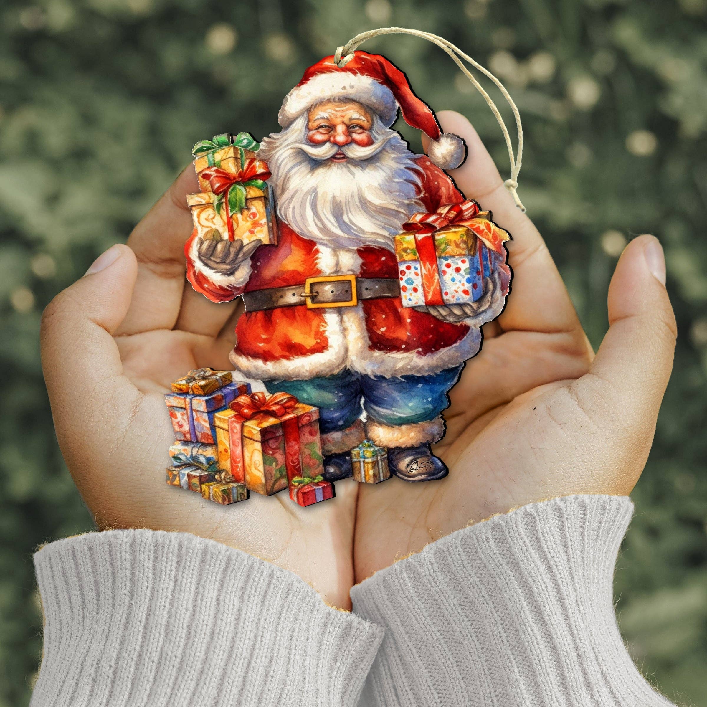 Hands holding the Merry Delivery Santa Wood Christmas Tree Ornament by artist g Debrekht, surrounded by colorful gift boxes, with a blurred green background.