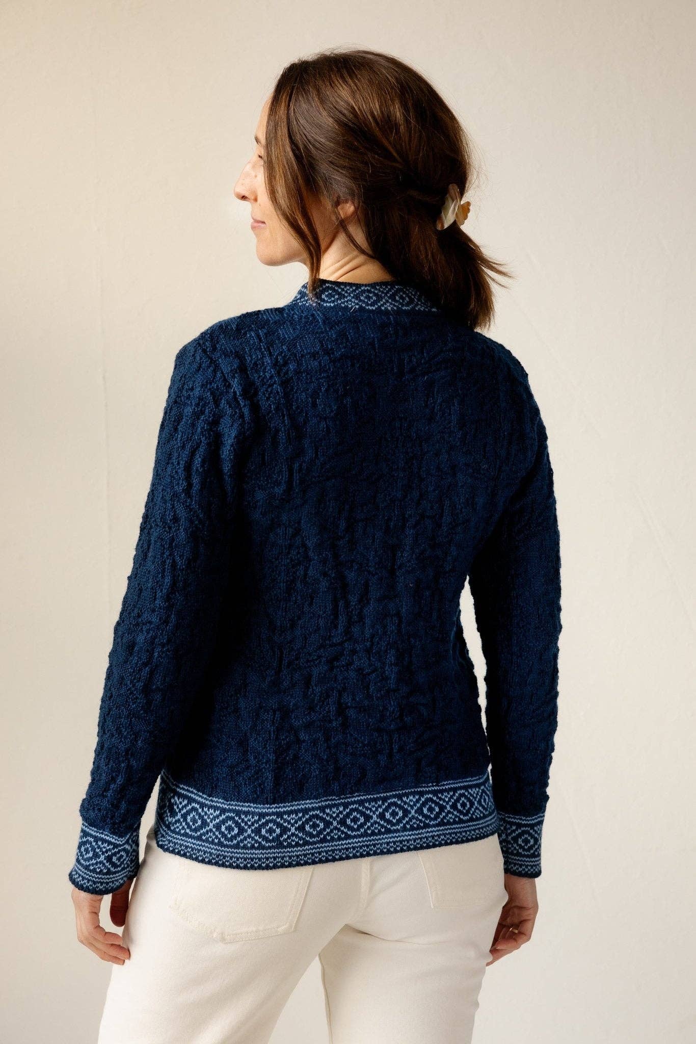 A woman with brown hair in a clip is wearing the Bryggen Women's Norwegian Cardigan in white and off-white pants, facing away from the camera.