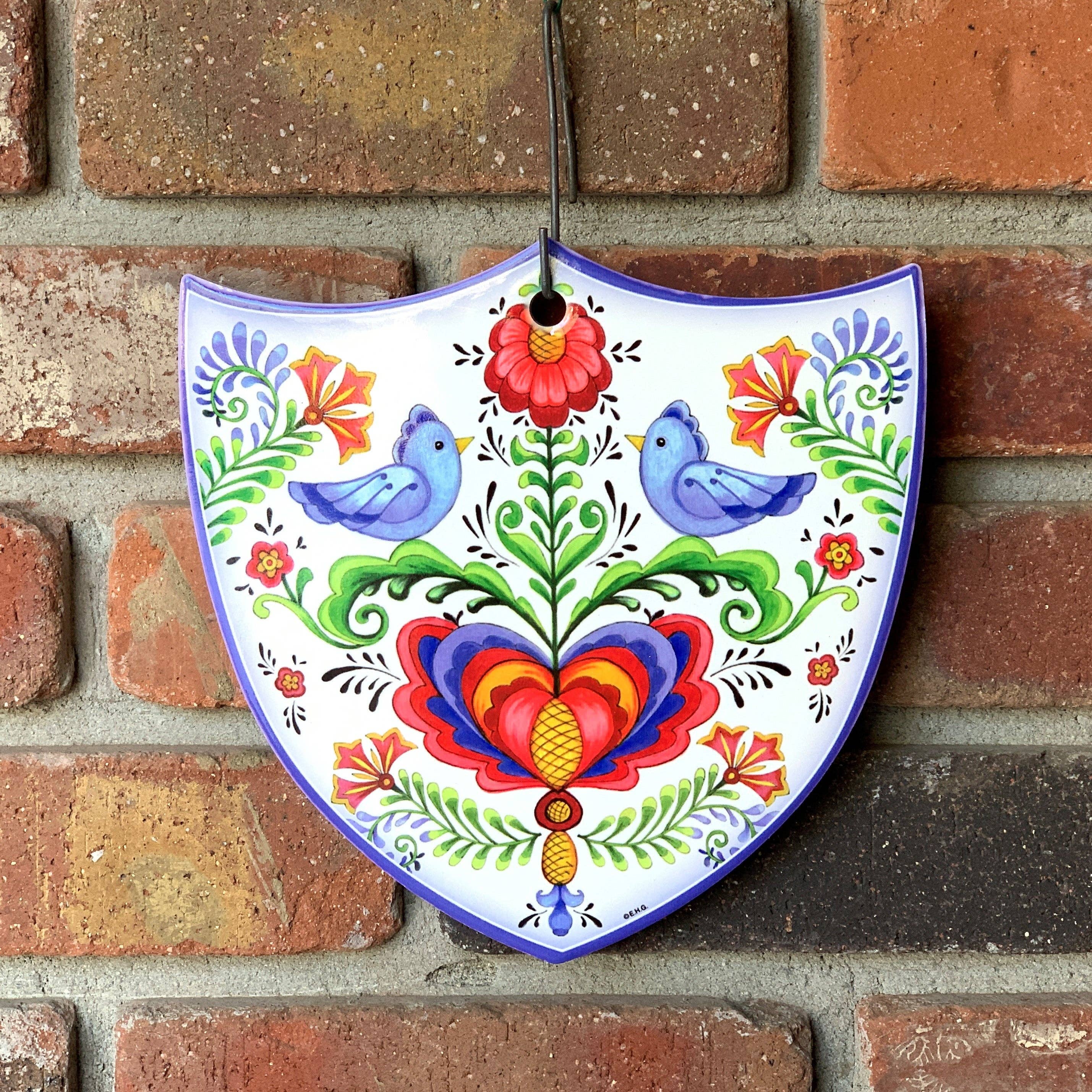 The Tile: Lovebirds Folk Art Cork Backed Ceramic Shield is a Nordic-designed wall decor featuring two blue birds, a heart, and flowers. This colorful ceramic plaque hangs beautifully on any wall.