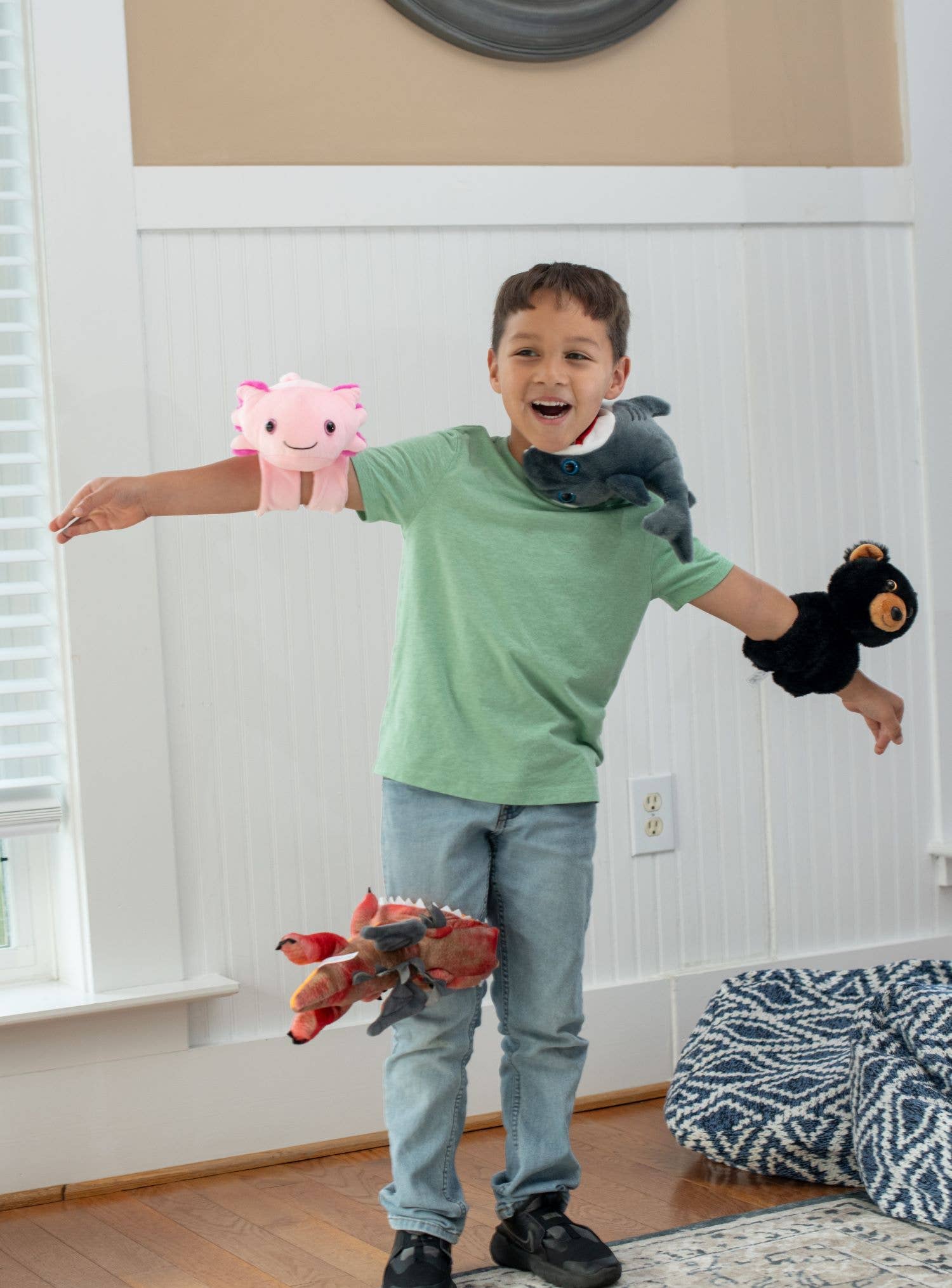 A boy in a green shirt and jeans stands indoors with plush toys on his arms, including the Plush: Sloth Slap Bracelet Slapstix (10 long, 7 body), while one stuffed animal is mid-air.