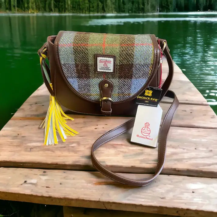 A Harris Tweed Saddle Bag in dark brown with blue, brown, and yellow tartan wool rests on a wooden dock by a green lake, its tag reflecting the handcrafted Scottish bag’s unique charm.