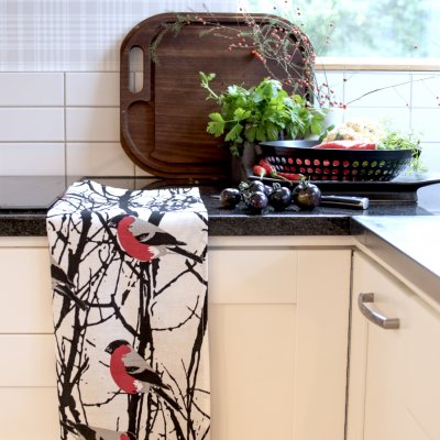 A kitchen counter with fresh herbs, veggies in a colander, and the Bullfinches in Birch Trees "Domherrar" tea towel by Almedahls of Sweden—red birds on white brighten this charming Swedish kitchen scene.