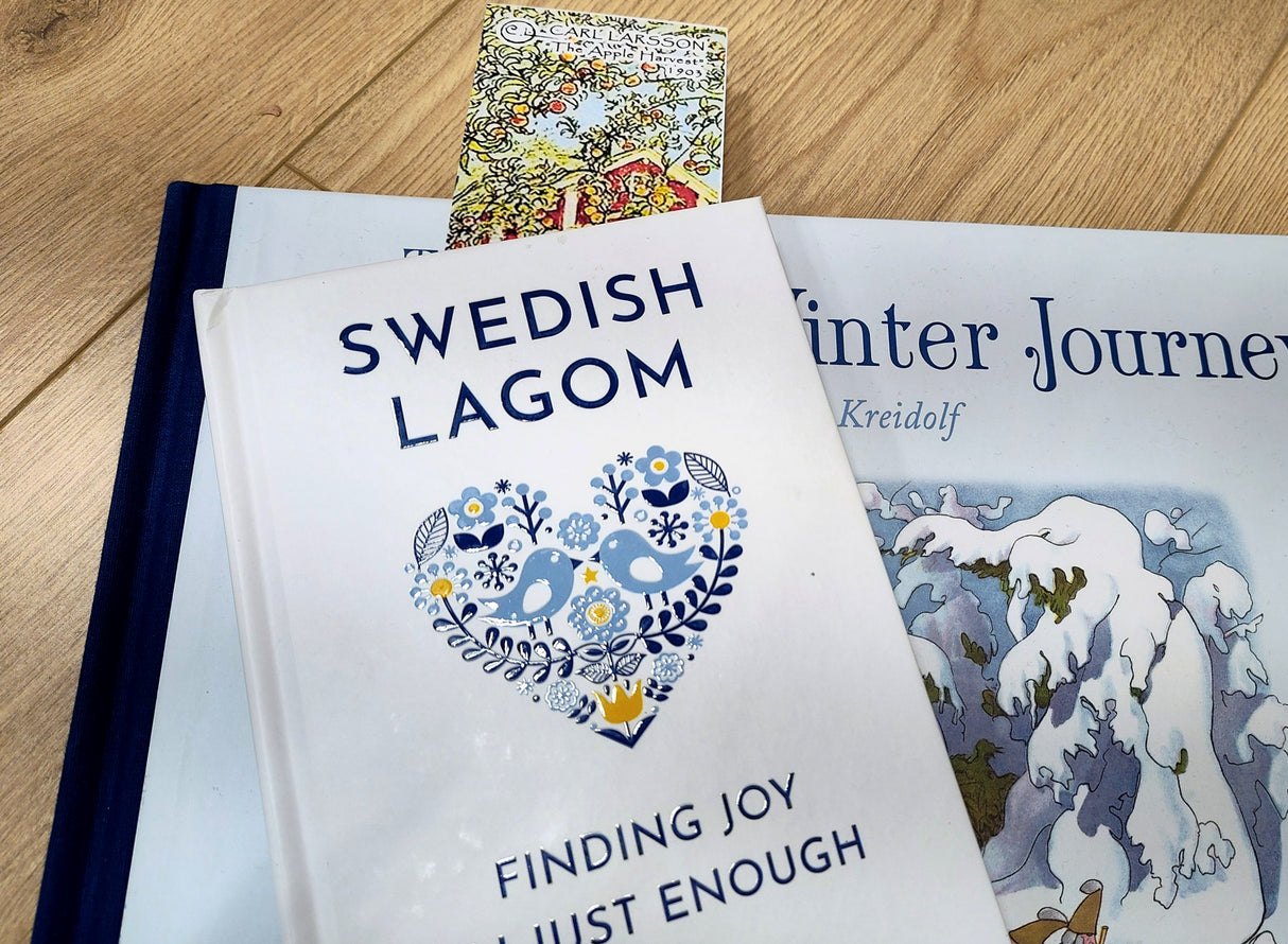 A book titled Swedish Lagom: Finding Joy in Just Enough rests atop another book and wood surface, with a postcard and the Bookmark: Carl Larsson 6-Piece Bookmark Collection (8.5" × 2.5" each) partially visible in the background.