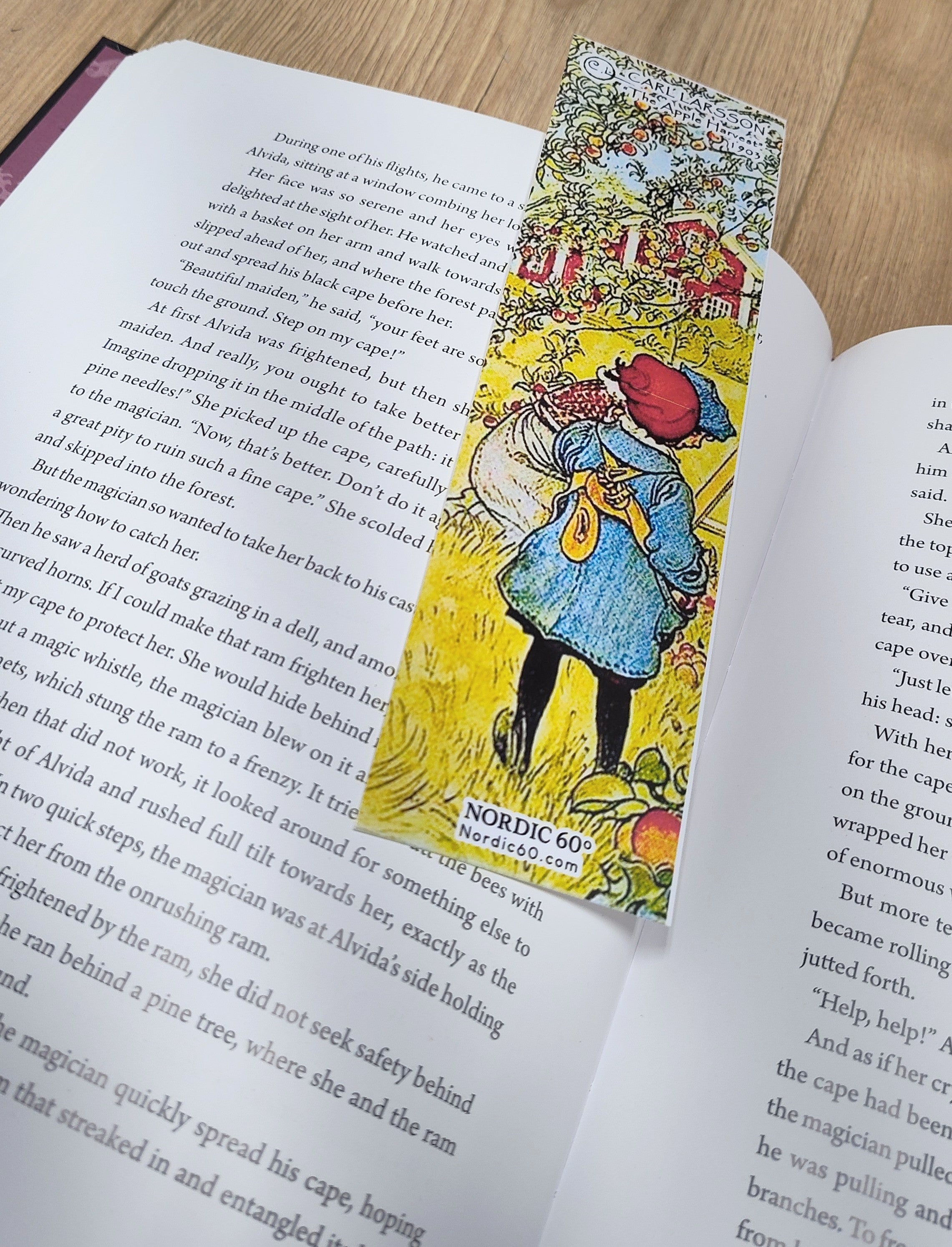 An open book rests on a wooden surface with the Carl Larsson “The Apple Harvest” bookmark (8.5" × 2.5") placed between its pages, adding an elegant touch of Scandinavian artistry.