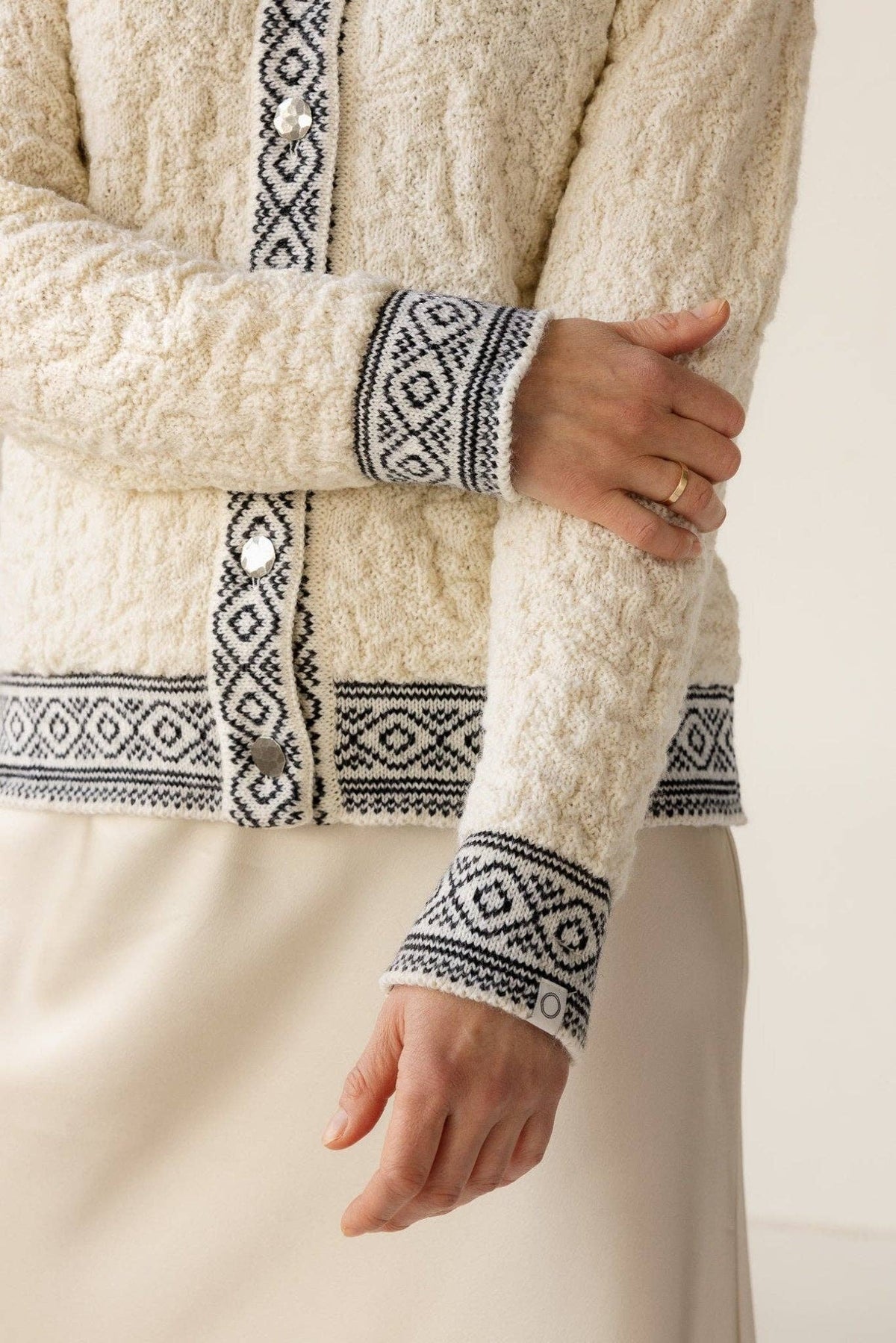 A person wearing the Bryggen Women's Norwegian Cardigan in white, featuring black geometric patterns on the cuffs and button band, paired with a cream skirt.