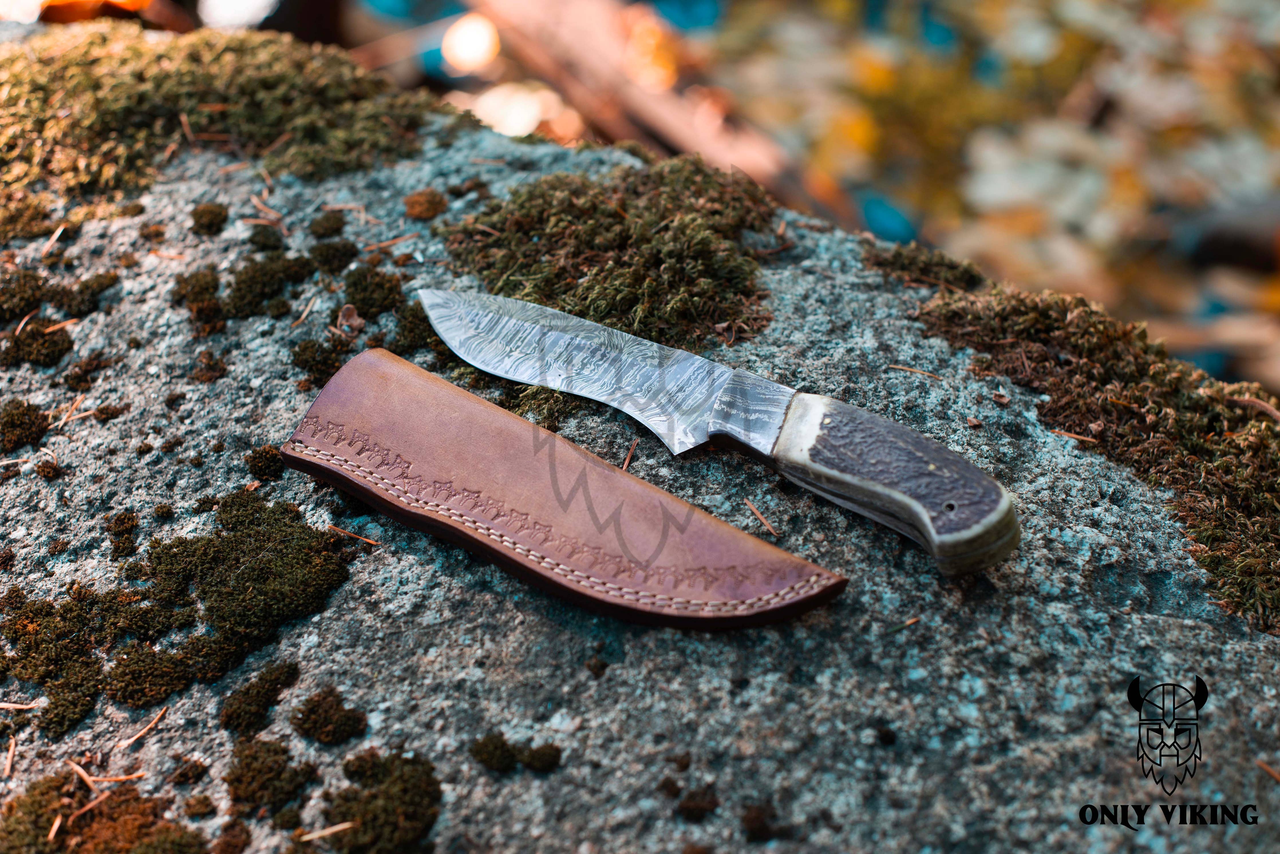 The Knife: Damascus Hunting Knife With Leather Sheath, featuring a patterned blade and textured handle, rests on a mossy rock, perfect for outdoor activities.