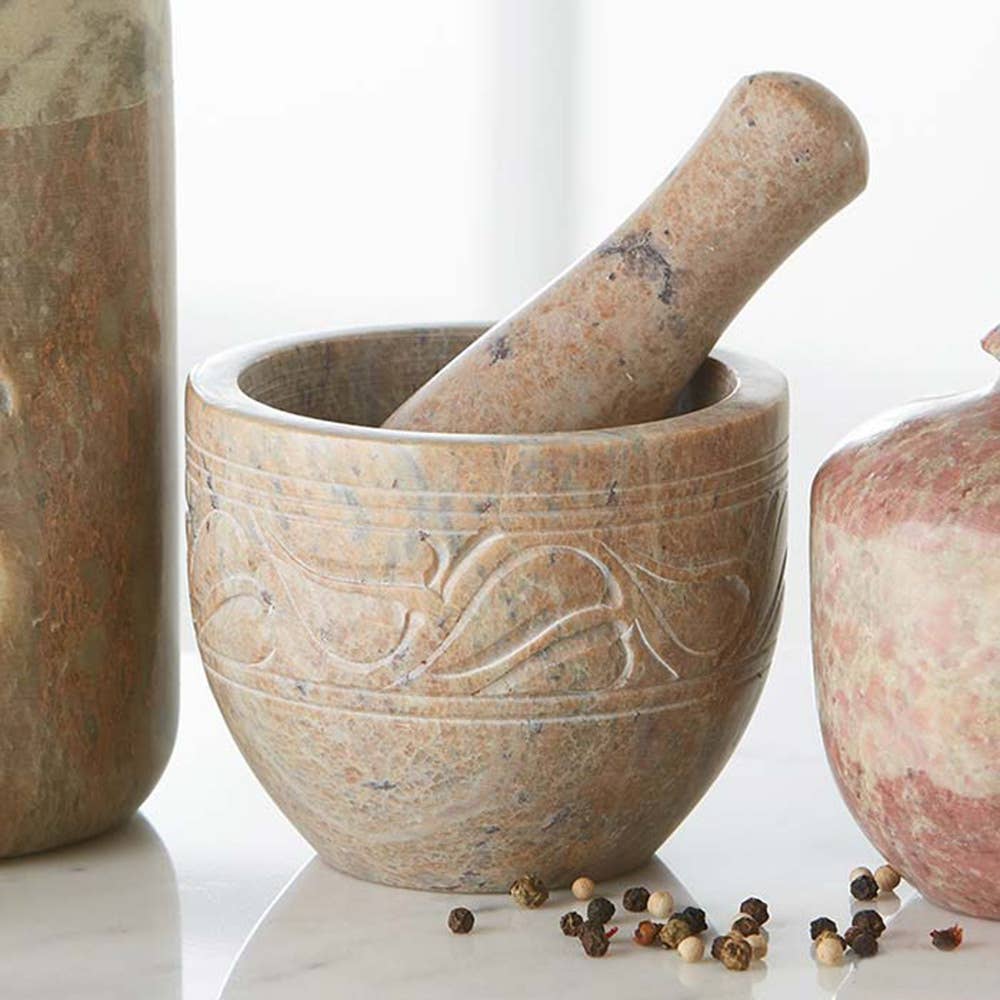 The Mortar & Pestle: Etched Gorara Rose Stone Mortar & Pestle features intricate patterns and rests on a countertop, surrounded by scattered peppercorns.