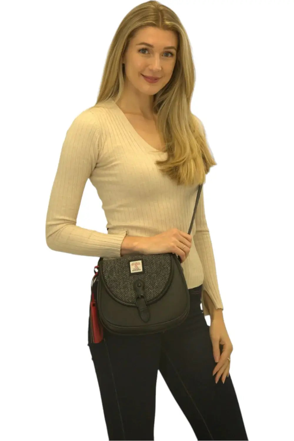 A woman with long blonde hair wears a beige sweater and dark jeans, holding a grey Harris Tweed saddle bag with a flap and red keychain.