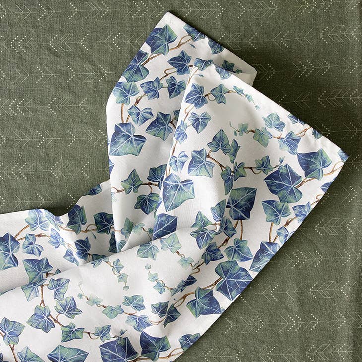 The Secret Garden Ivy Organic Tea Towel by Koustrup & Co. features white organic cotton with a blue and green ivy pattern over dark green fabric, boasting a subtle dotted and arrow design, reflecting Koustrup & Cos dedication to elegance and sustainability.