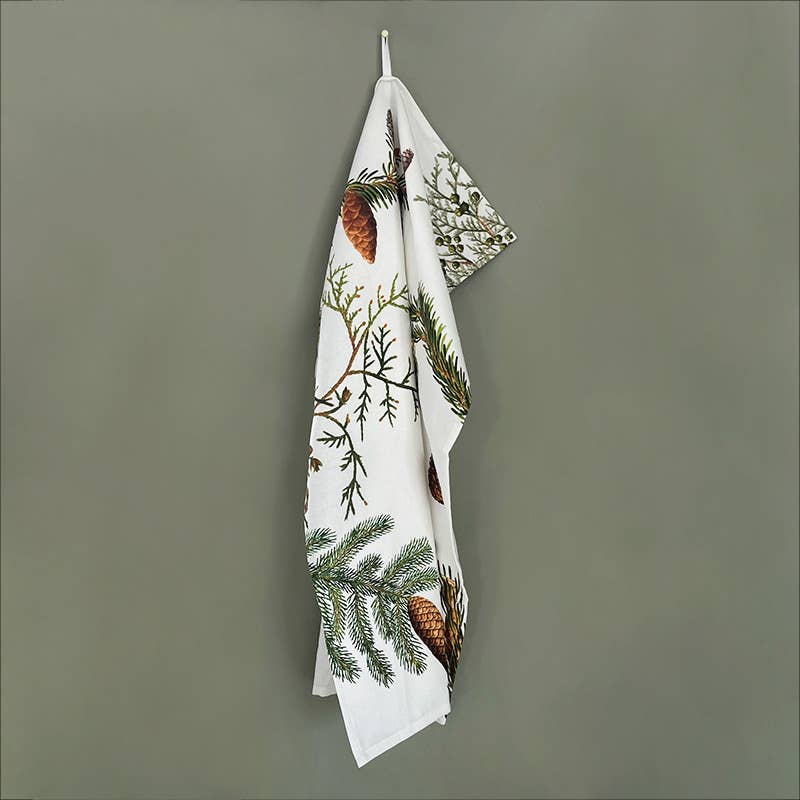 Displayed on a hook against a gray wall, the "Tea Towel: Pine Trees" from Koustrupco showcases an elegant design of pine cones and branches. Made from 100% organic cotton, it brings the peaceful essence of pine trees into your kitchen. Ideal for eco-friendly households, it is packaged in biodegradable materials.