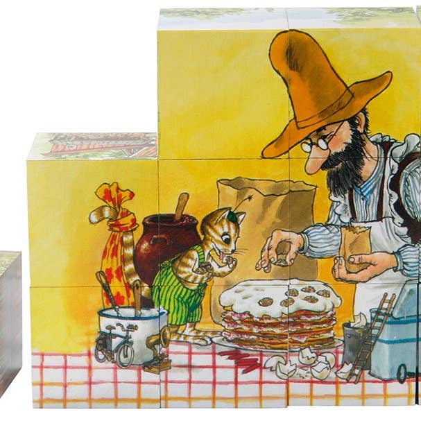 An illustration showing a man in a hat with a small cat, capturing the charm of Pettson & Findus, as they assemble a layered cake on a checkered tablecloth. Wooden blocks are scattered around, enhancing the whimsical atmosphere. This scene is available as part of the Puzzle: Pettson & Findus, 6-Pack (12 Pieces).