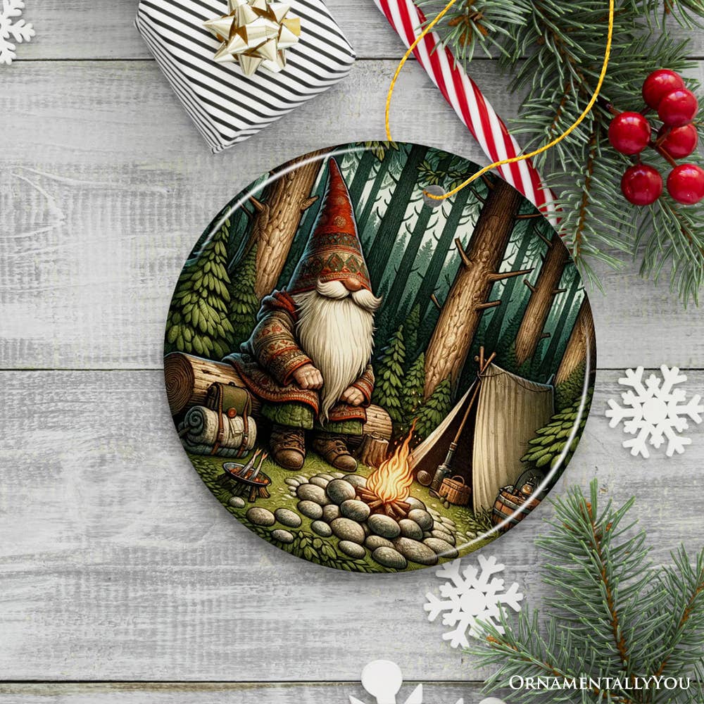 The Nordic Folk Gnome Forest Elder Themed Ceramic Ornament features a gnome by a campfire among snowflakes, pine branches, and a gift box. Embrace the cozy Scandinavian spirit with this charming decoration in your festive collection.