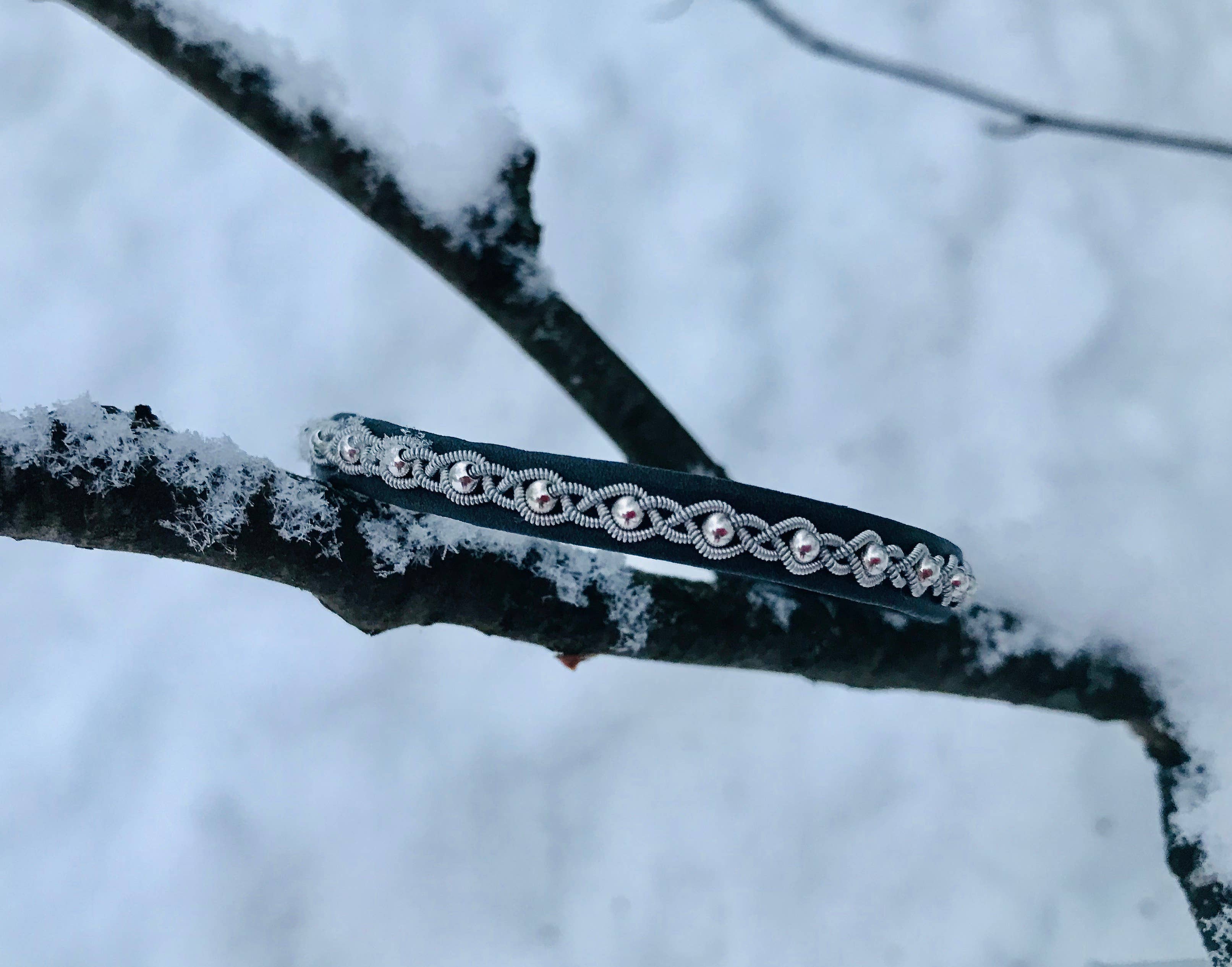 The Bracelet: Sami Lapland Bracelet With Reindeer Hide Pewter & Silver Beads #27: Black rests on a snow-covered tree branch, its silver and black design crafted from reindeer hide and accented with pewter beads, set against a snowy backdrop.