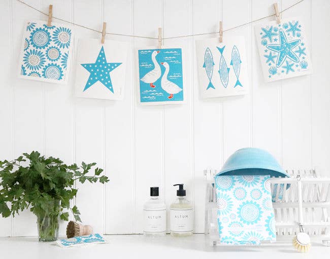 Cards with blue designs hang on a string above a white shelf, alongside soap dispensers, a green plant, biodegradable cleaning supplies, and an absorbent Swedish dish cloth featuring a turquoise fish design.